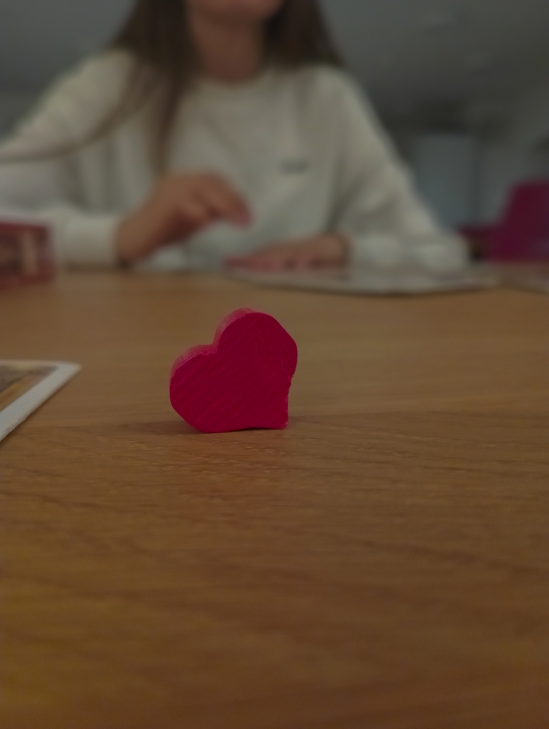 Nahaufnahme eines kleines hölzernen Herzes auf einem Tisch, im Hintergrund eine verschwommene Person